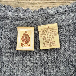 May include: Close-up of a gray knitted sweater with a textured pattern. The sweater has a scalloped neckline. Two tags are visible, one with the brand name "Henna" and the other with care instructions and material composition.