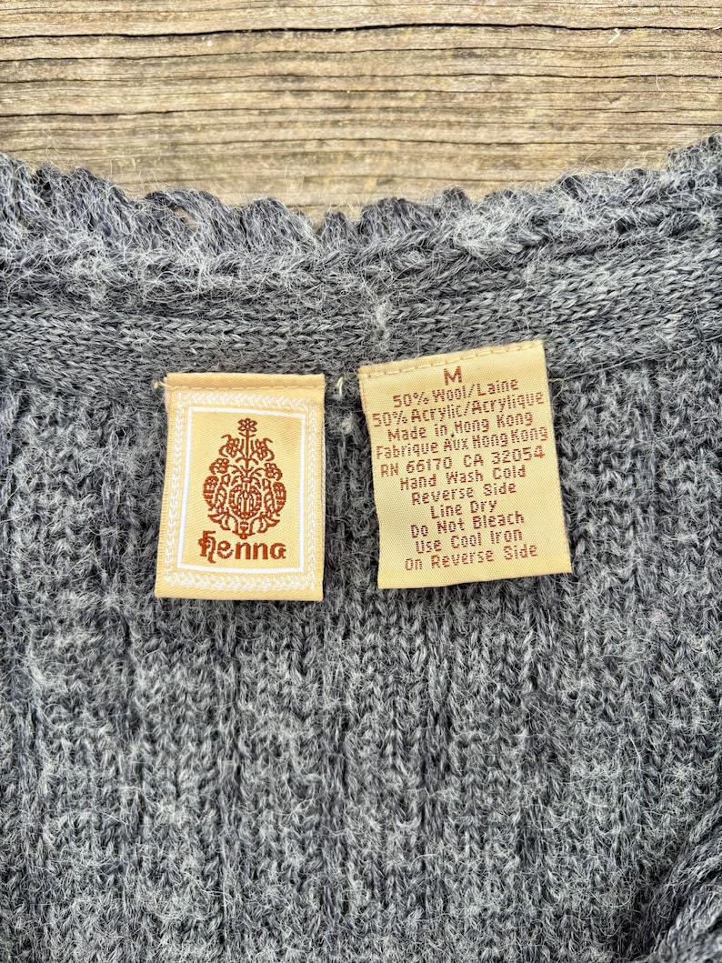 May include: Close-up of a gray knitted sweater with a textured pattern. The sweater has a scalloped neckline. Two tags are visible, one with the brand name "Henna" and the other with care instructions and material composition.