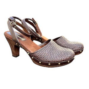 May include: Brown leather clogs with a woven texture on the toe box and a wooden heel. The shoes have an ankle strap with a buckle and are adorned with small gold-colored studs around the sole.