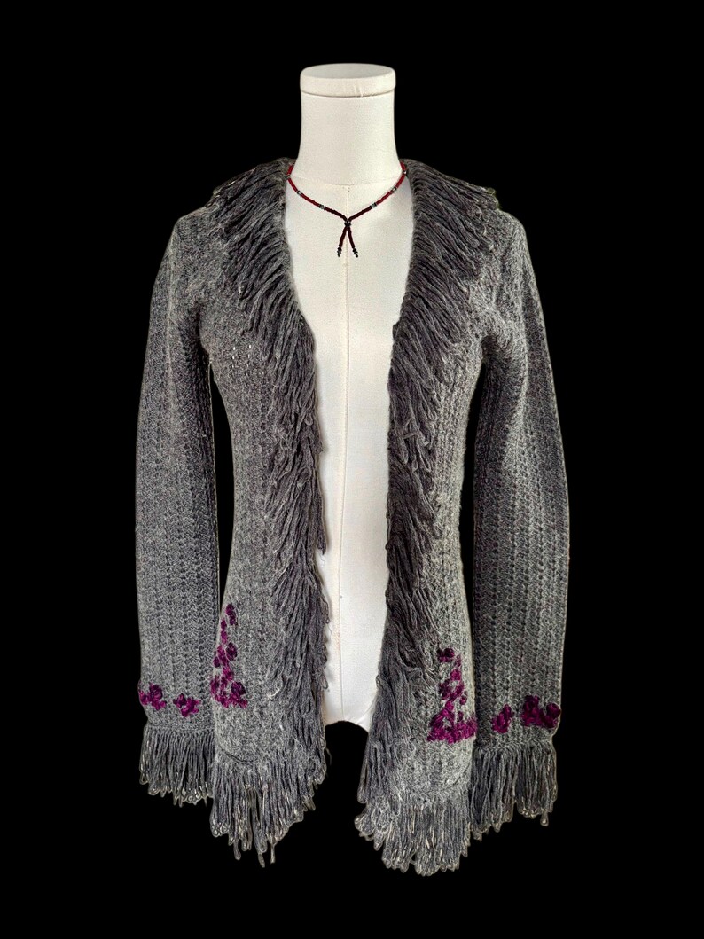 May include: A gray, open-front cardigan with fringe trim and purple floral embroidery. The long-sleeved sweater is displayed on a mannequin against a black background. A beaded necklace is also visible.