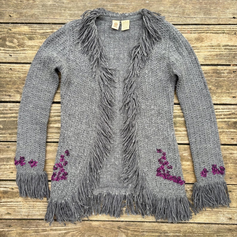 May include: A gray knit cardigan with fringe detailing along the front, collar, and cuffs. The cardigan features embroidered purple floral accents near the cuffs and hem. The open-front design and long sleeves create a bohemian style.