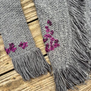 May include: A gray knitted scarf with fringe and embroidered magenta floral accents. The scarf is made of a soft, textured material and has a detailed knit pattern. The fringe adds a decorative element to the scarf.