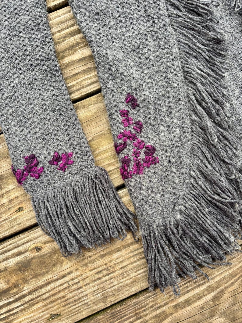 May include: A gray knitted scarf with fringe and embroidered magenta floral accents. The scarf is made of a soft, textured material and has a detailed knit pattern. The fringe adds a decorative element to the scarf.