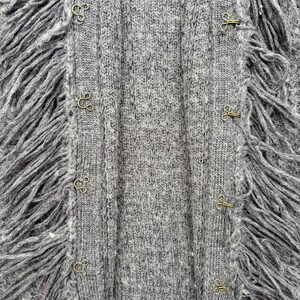 May include: Close-up of a gray knit cardigan with fringe trim. The cardigan features a ribbed texture and is fastened with gold-colored hook-and-eye closures. The fringe adds a textural element to the garment.