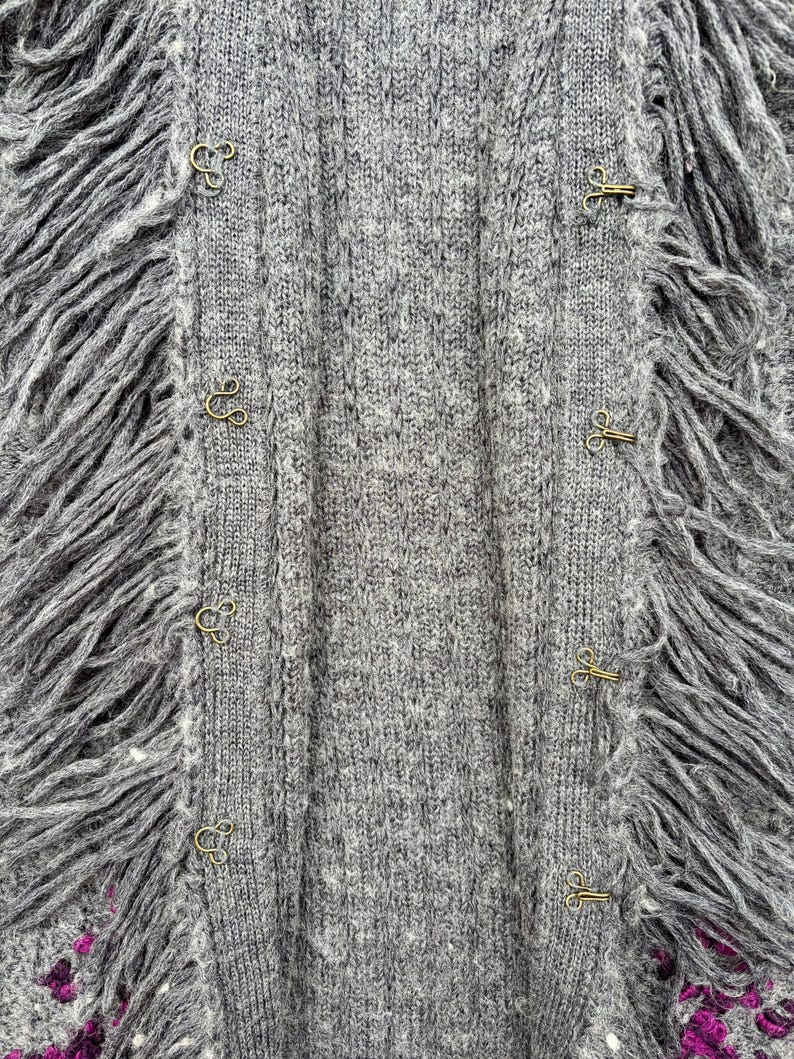 May include: Close-up of a gray knit cardigan with fringe trim. The cardigan features a ribbed texture and is fastened with gold-colored hook-and-eye closures. The fringe adds a textural element to the garment.