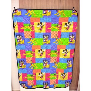 Puede incluir: Una manta acolchada con un patrón repetitivo de Mickey Mouse y Pluto sobre un fondo de patchwork colorido de cuadrados azules, verdes, naranjas y rosas con varias texturas y puntos, terminada con un borde de tela de color negro sólido.