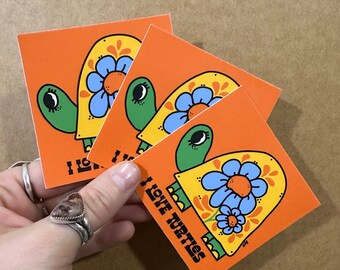 May include: Three orange square stickers with a cartoon turtle on each. The turtles are green with blue flowers and yellow accents. The text "I love turtles" is written on one of the stickers.