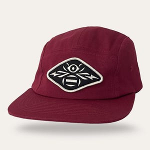 May include: A maroon five-panel cap featuring a diamond-shaped patch on the front. The patch is black with a white embroidered graphic of an eye above a stylized bee or insect shape, outlined in white thread, with a flat brim and stitched seams.