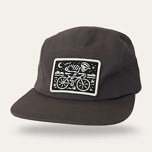 May include: A dark gray five-panel cap featuring a rectangular patch on the front. The patch displays a white embroidered graphic of a skeleton riding a bicycle under a crescent moon and stars against a black background with a white border.