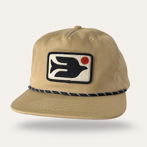 May include: A tan baseball cap featuring a rectangular patch on the front. The patch displays a black bird silhouette and a small red circle on a cream background. A black and white speckled rope trim runs across the base of the front crown.