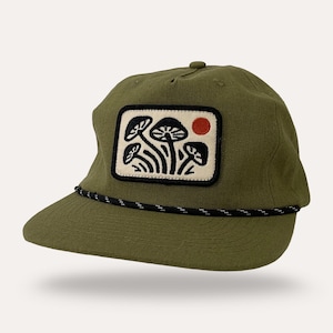 May include: Olive green baseball cap with a black and white rope detail. The cap features a rectangular patch with a mushroom design and a red sun. The cap is made of a durable fabric.