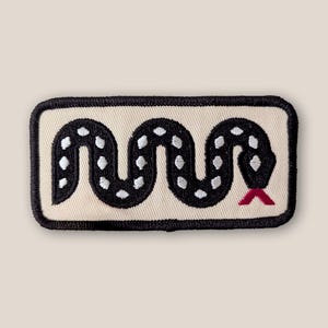 May include: A rectangular embroidered patch featuring a black snake with white spots arranged in a wavy, horizontal pattern on a cream-colored background. The snake has a small, pointed red tongue extending from its head, and the entire patch is bordered by a thick, black stitched edge.