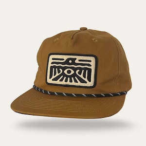 May include: A brown baseball cap with a black and white rope detail on the brim. The cap features a rectangular patch with a black and cream-colored graphic design. The design includes a bird-like figure and geometric shapes.