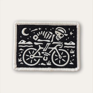 May include: A rectangular embroidered patch featuring a black background with white stitching. The design depicts a skeleton wearing a cap while riding a bicycle under a crescent moon and stars, with simple landscape elements at the bottom.