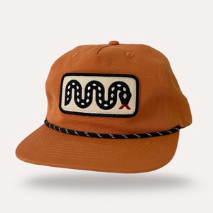 May include: A burnt orange baseball cap with a black and white rope brim. The cap features a rectangular patch with a black and white snake design and a red tongue.