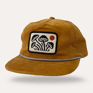 May include: Mustard yellow corduroy baseball cap with a patch featuring a black and white mushroom design and a red circle. The cap has a curved brim and a black and white rope detail along the brim.