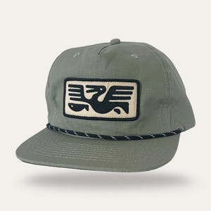 May include: A sage green baseball cap with a black and white rope detail along the brim. The cap features a rectangular patch with a black bird design on an off-white background.