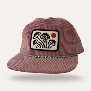 May include: A dusty rose corduroy cap with a curved brim. The cap features a rectangular patch with a black and white mushroom design and a red sun. A black and white rope detail accents the brim.
