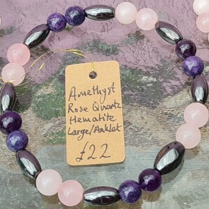 Może przedstawiać: Bransoletka lub anklet z koralików z ametystu, kwarcu różowego i hematytu. Projekt zawiera fioletowe, różowe i szare koraliki w okrągłych i owalnych kształtach. Mała etykieta zawiera szczegółowe informacje o materiałach i rozmiarze.