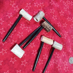 May include: A collection of small, handcrafted hammers with black handles and antler heads. The hammer heads are various shades of white, brown, and tan. The hammers are arranged on a red fabric with white star patterns.