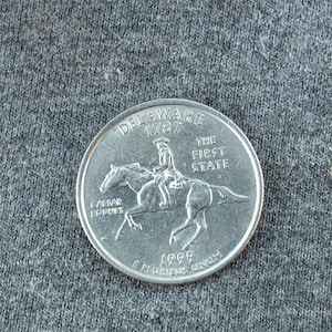 May include: A silver Delaware quarter coin featuring Caesar Rodney on horseback, with the text "DELAWARE 1787 THE FIRST STATE" and the year 1999. The coin is on a gray surface.