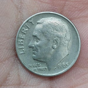 May include: A close-up of a silver 1965 dime. The coin features a profile of a man, the word "LIBERTY" at the top, and the phrase "IN GOD WE TRUST" at the bottom. The year "1965" is also visible.