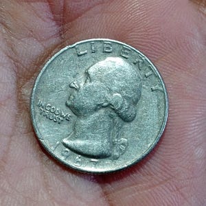 May include: A silver quarter coin from 1967, featuring a profile of George Washington and the inscriptions "LIBERTY" and "IN GOD WE TRUST." The coin is held in a hand, showing signs of wear.