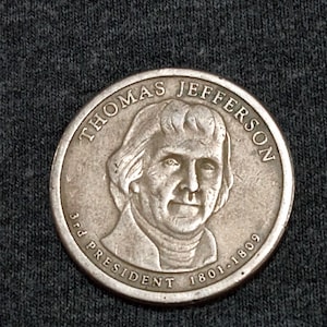May include: A silver coin featuring a portrait of Thomas Jefferson, with the text "THOMAS JEFFERSON" above the portrait. The coin also includes the text "3rd PRESIDENT 1801-1809".