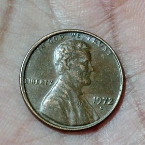 May include: A copper-colored United States penny from 1972, featuring a profile of Abraham Lincoln. The coin has the inscriptions "IN GOD WE TRUST" and "LIBERTY".