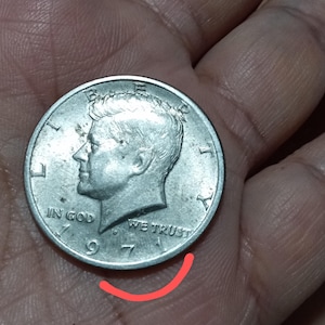 May include: A silver 1971 Kennedy half dollar coin. The coin features a profile of John F. Kennedy and the inscriptions "LIBERTY", "IN GOD WE TRUST", and "1971". The coin is held in a hand.