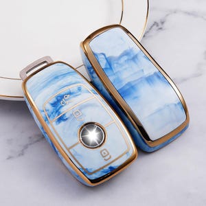 May include: Two car key fobs with a blue and white marbled design and gold trim. The key fobs have a metallic silver key ring and a gold border. The key fobs have a button in the center.