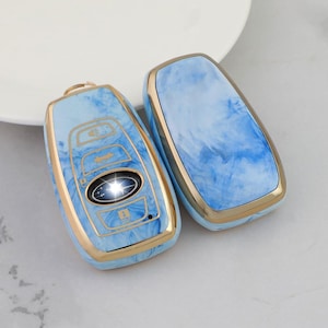 May include: Two car key fobs with a light blue and white marbled design. The fobs have a gold-colored trim. One fob has buttons and a Subaru logo. The other fob is plain. The fobs are on a white surface.