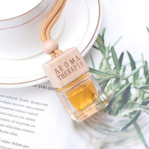 May include: A wooden car air freshener with a brown cord. The air freshener has a small glass bottle filled with a yellow liquid and the words "Aroma Therapie" engraved on the wood.