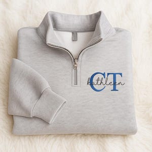 Peut inclure: Un sweat-shirt gris clair avec un col zippé quart de tour. Le sweat-shirt porte le nom brodé "Kathleen" en écriture cursive, avec les initiales "CT" en bleu. Le sweat-shirt est plié sur une surface blanche et moelleuse.