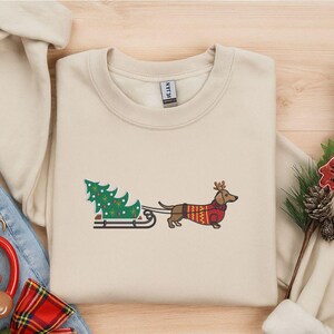 May include: A cream-colored sweatshirt featuring an embroidered Christmas design. The design includes a dachshund wearing a reindeer antler headband and a red sweater, pulling a Christmas tree on a sled. A festive holiday theme.