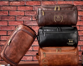 May include: Four brown leather toiletry bags stacked on top of each other. The top bag has the name "Wesley" and the date "Est. 2023" embossed on it. The second bag has the name "Michael" and the text "Groomsman 12.16.2020" embossed on it. The third bag has the initials "A&J" and the text "Anna & John" embossed on it.