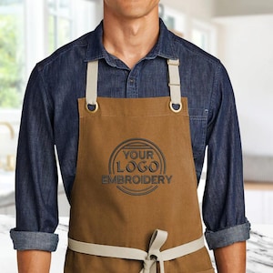 Op de afbeelding: Een bruine canvas schort met verstelbare banden en een grote cirkel geborduurd met de tekst "YOUR LOGO EMBROIDERY".