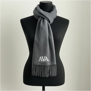 May include: A gray scarf with fringe detail and the embroidered initials "AVA" in white. The scarf is draped around a black mannequin, showcasing its texture and design. A stylish accessory for any wardrobe.