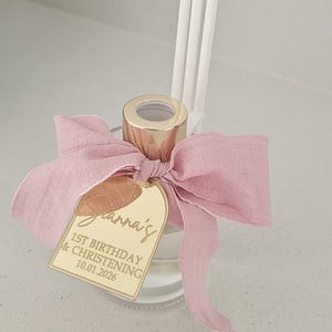 May include: A clear glass diffuser bottle with a gold-coloured cap, a pink ribbon, and a gold tag. The tag reads "Jianna's 1st Birthday & Christening 10.01.2026". White diffuser reeds are inserted into the bottle.