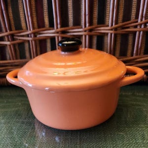 May include: A small orange ceramic pot with a black knob lid and two handles. The pot is sitting on a green surface.