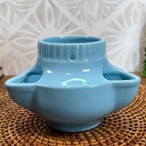 May include: A light blue ceramic planter with a unique design. The planter has a wide, flat base and a rounded top with a hole in the center. It has three small openings on the sides, perfect for holding small plants or succulents.