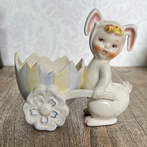May include: A ceramic Easter bunny figurine pulling a pastel egg-shaped cart. The bunny is white with pink accents and a sweet expression. The cart has a flower wheel and is painted in soft yellow and blue.