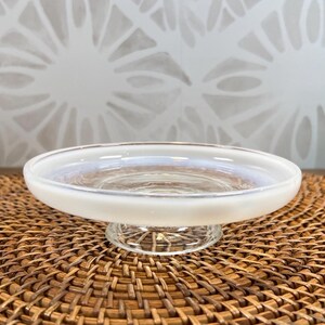 May include: A clear glass pedestal dish with a frosted white rim. The dish sits on a clear, textured base. The dish is on a brown woven mat, with a white patterned background.