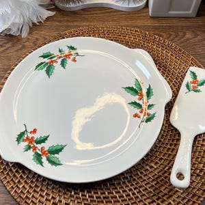 May include: A white ceramic serving plate and matching server with a holly berry design. The plate has handles and the server has a handle with a hole. The set is on a woven placemat.