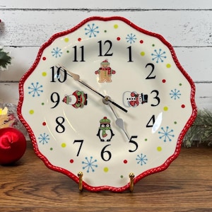 May include: A decorative Christmas-themed clock. The clock face is a white plate with a red scalloped edge. It features illustrations of a gingerbread man, snowman, and penguin. The clock hands are black.