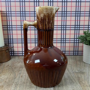 May include: A brown ceramic pitcher with a glazed finish. The pitcher has a wide base and a narrow neck with a pouring spout. The pitcher has a light brown glaze on the top and a darker brown glaze on the bottom.