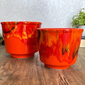 May include: Two ceramic planters with a vibrant, glossy finish. The larger planter is on the left, and the smaller one is on the right. Both have a bright orange base with a fiery, abstract pattern of red, yellow, and black. The planters are suitable for indoor or outdoor use.