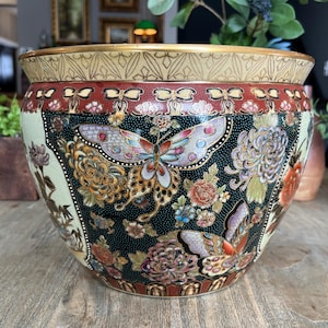 May include: A decorative ceramic planter with a wide, rounded shape. The planter features a cream-colored base with a black central band adorned with butterflies and floral patterns in shades of red, orange, and gold. The rim has a gold geometric design.