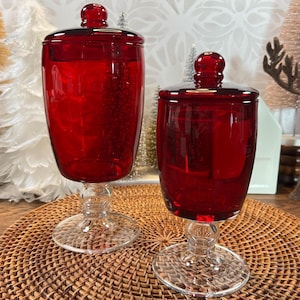May include: Two ruby red glass apothecary jars with lids. The jars have a bulbous shape, clear glass stems, and round bases. The lids are topped with a small, round knob. The jars are displayed on a woven, brown placemat.