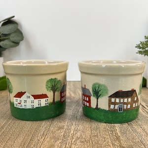 May include: Two small ceramic cream-colored pots with green, red, and white painted scenes of houses and trees. The pots are approximately 4 inches tall.